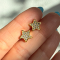 Gold star-shaped earrings with clear stones held between fingers against a blurred background