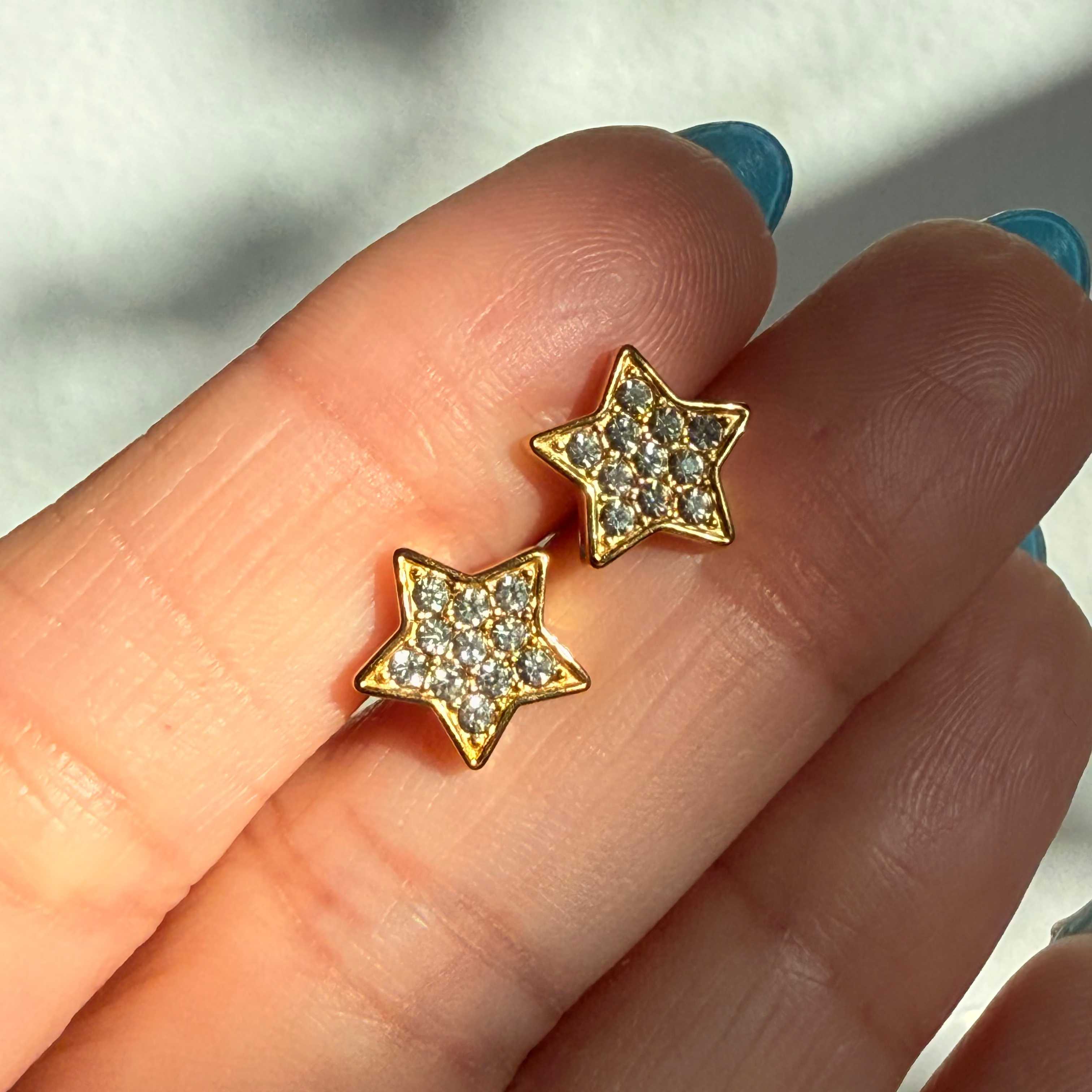 Gold star-shaped earrings with clear stones held between fingers against a blurred background